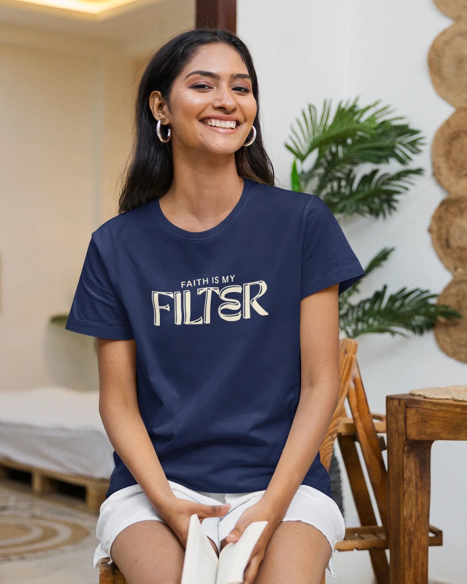 A woman sitting on a wooden chair wearing a navy blue t-shirt with the text 'FAITH IS MY FILTER' in white, paired with white shorts and socks in a warm, modern indoor setting.