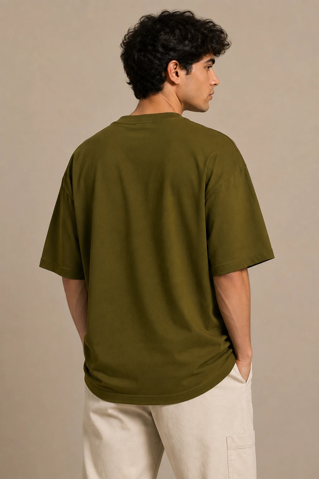 A man wearing an oversized olive green t-shirt and off-white pants, shown from the back against a neutral beige studio background.
