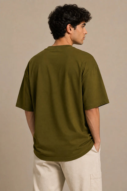 A man wearing an oversized olive green t-shirt and off-white pants, shown from the back against a neutral beige studio background.