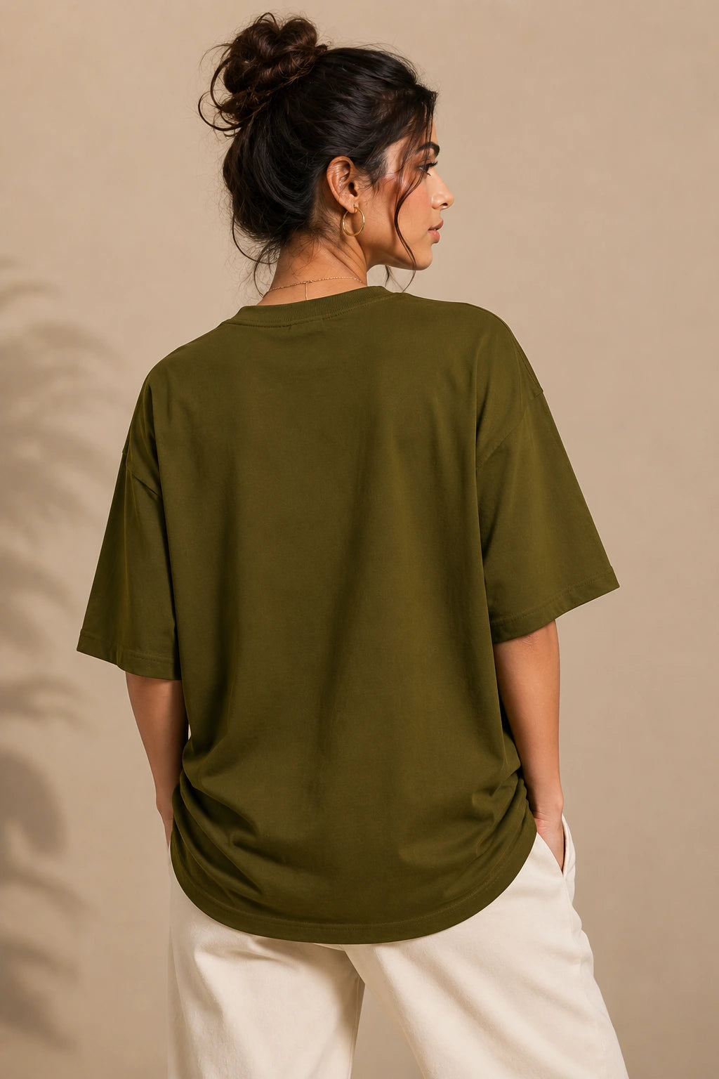 A woman shown from the back wearing an oversized olive green short-sleeve t-shirt and cream-colored pants, standing against a warm beige background.