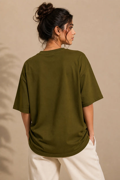 A woman shown from the back wearing an oversized olive green short-sleeve t-shirt and cream-colored pants, standing against a warm beige background.