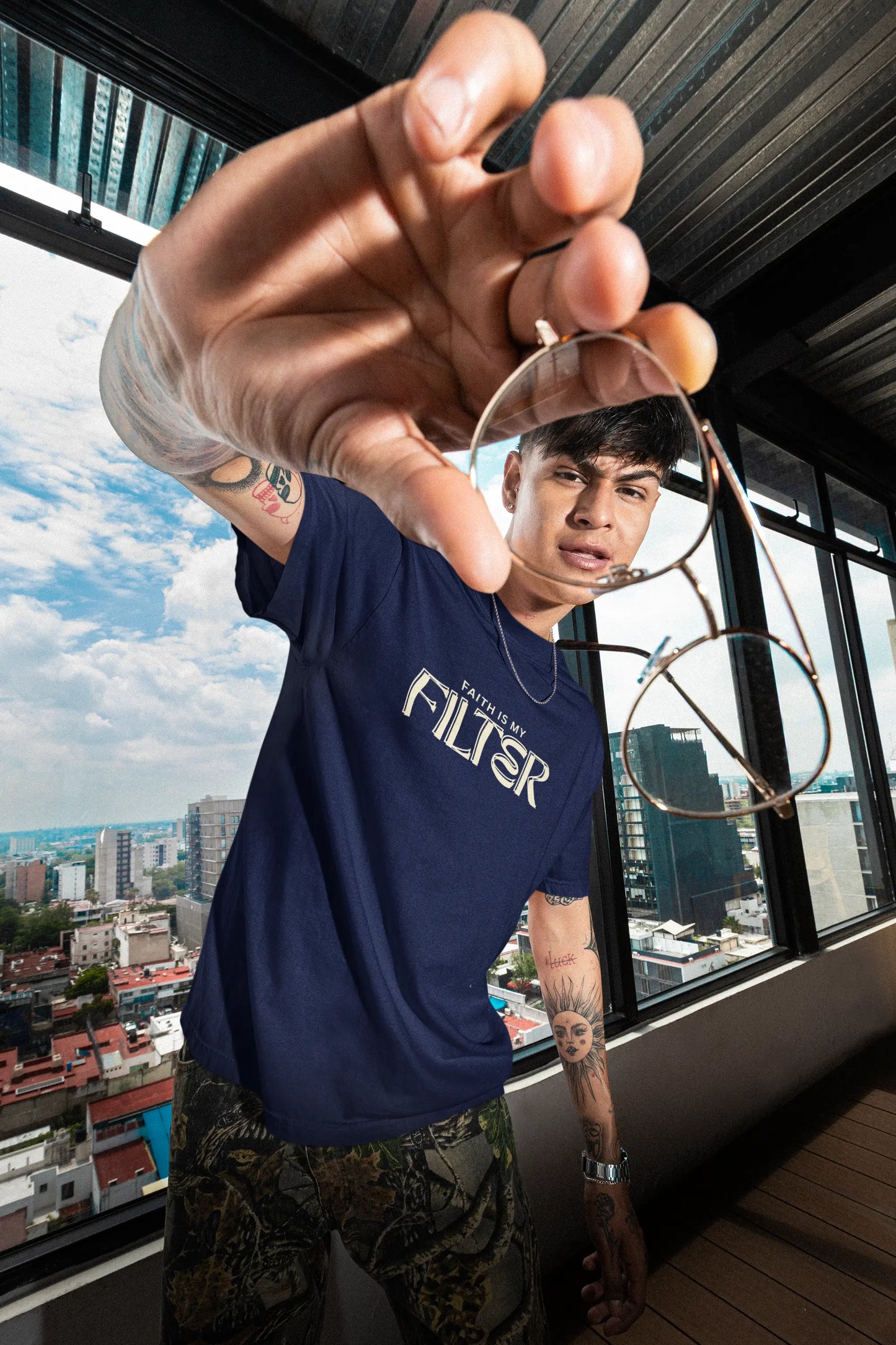 A man wearing a navy blue t-shirt with 'FILTER' printed in white across the chest, holding a pair of round metal-framed glasses in front of his face. He is standing in a modern room with large windows overlooking a city skyline.