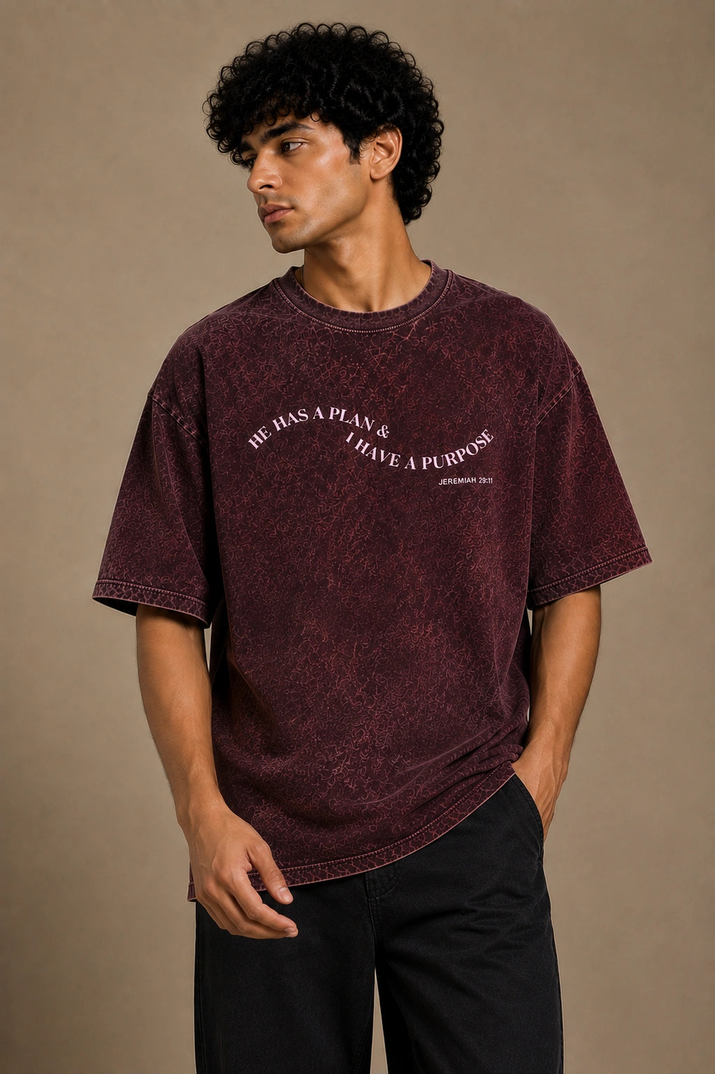 A man wearing a washed dark burgundy t-shirt with the text 'HE HAS A PLAN & I HAVE A PURPOSE' printed in white on the chest, paired with black pants, against a solid beige background.