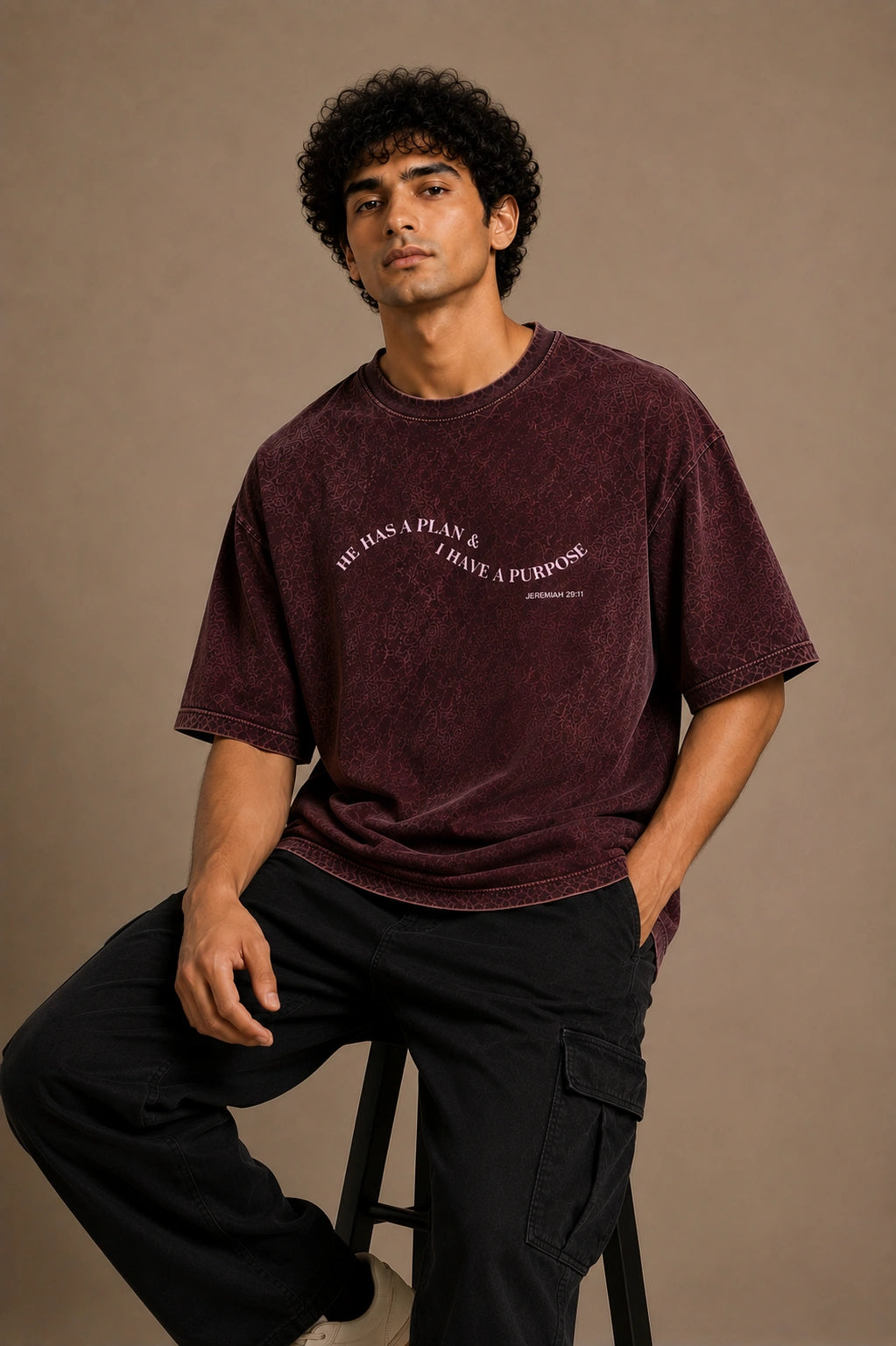 A man wearing a washed dark burgundy t-shirt with white text and black cargo pants, sitting on a stool against a solid beige background.