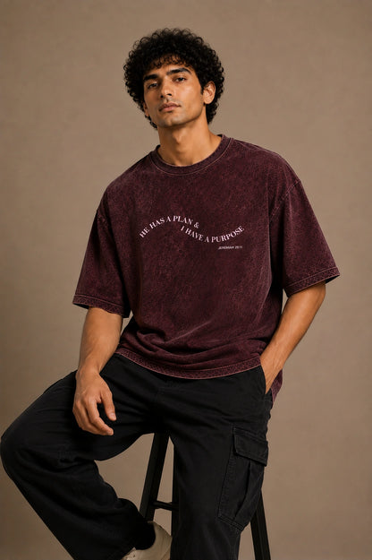 A man wearing a washed dark burgundy t-shirt with white text and black cargo pants, sitting on a stool against a solid beige background.