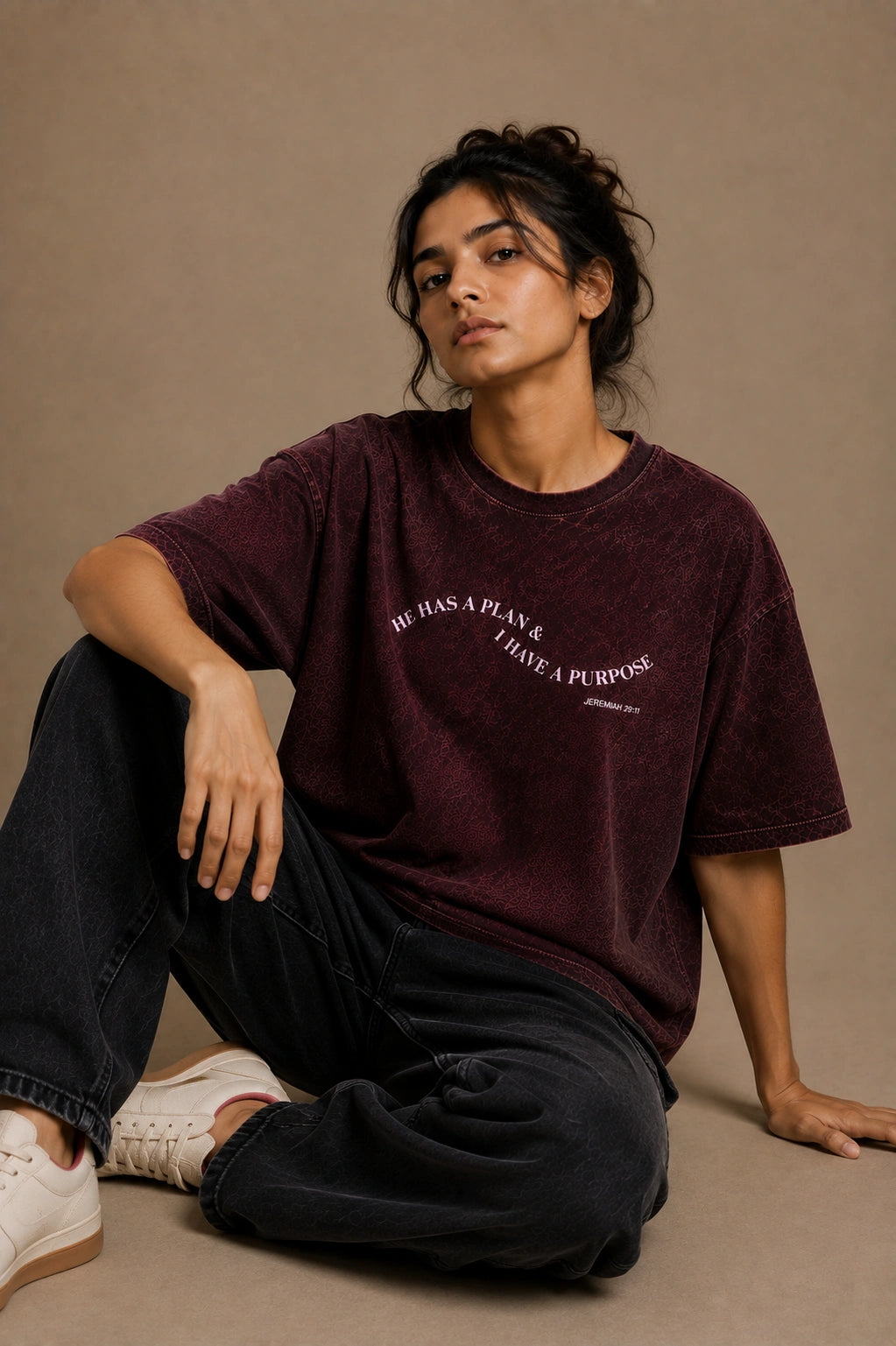 A model sits on the floor wearing a washed dark burgundy t-shirt with the text 'hE HAS A PLAN. I HAVE A PURPOSE.