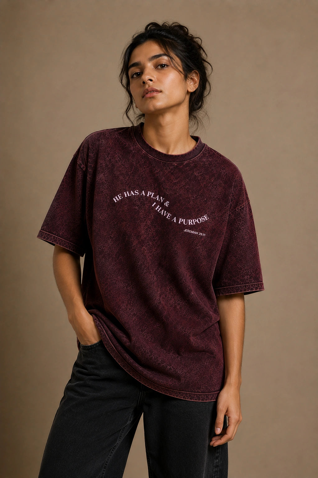 A woman wearing an oversized mineral-wash dark burgundy t-shirt with white text that reads 'HE HAS A PLAN & I HAVE A PURPOSE' and black jeans, posing against a solid beige studio backdrop.