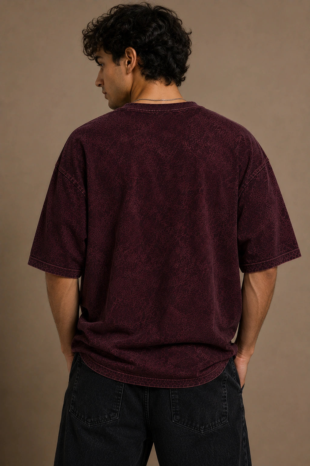A man wearing an oversized, washed dark burgundy t-shirt, shown from the back against a solid warm brown studio background.