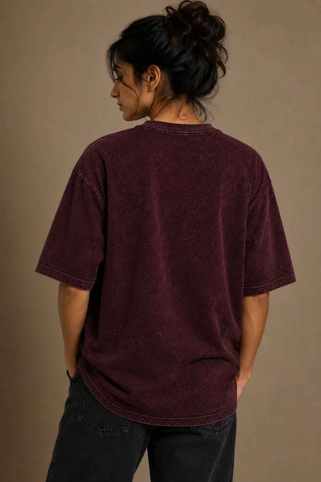 A person shown from the back wearing an oversized short-sleeve crewneck t-shirt in a washed dark burgundy or plum color, paired with dark jeans, against a plain warm taupe studio background.