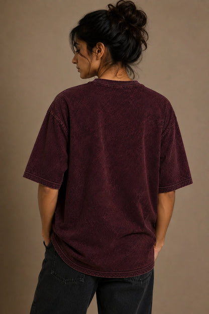 A person shown from the back wearing an oversized short-sleeve crewneck t-shirt in a washed dark burgundy or plum color, paired with dark jeans, against a plain warm taupe studio background.