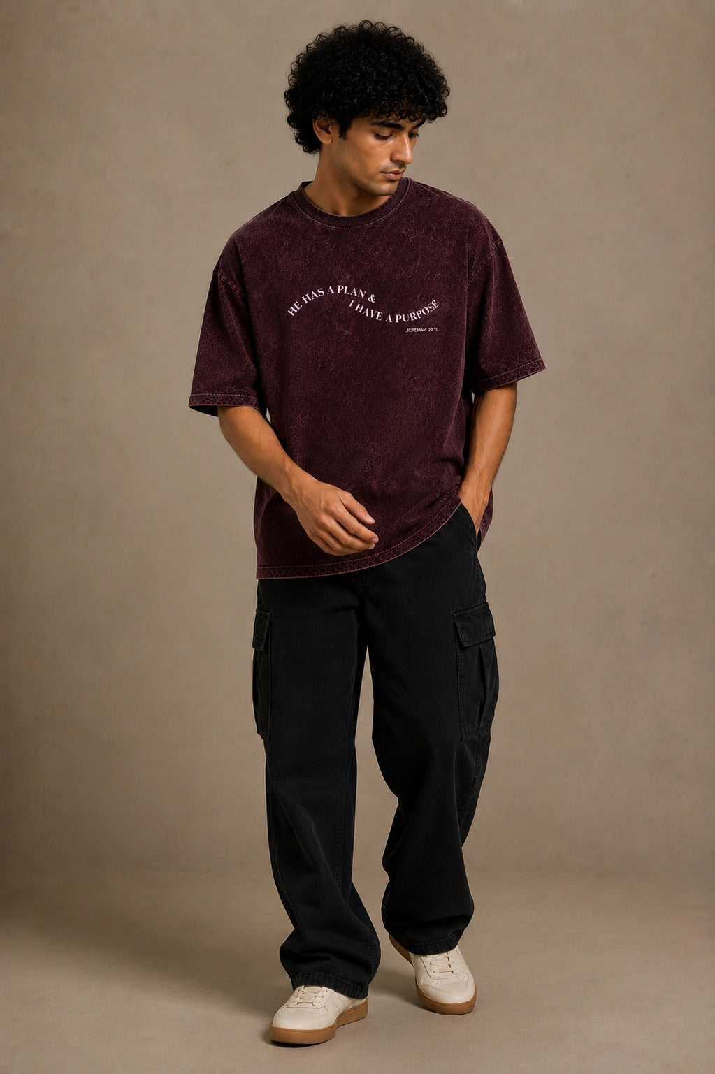 A man wearing a washed dark burgundy t-shirt with 'SANTO SEGUIN' printed on the chest, paired with black cargo pants and white sneakers, standing against a neutral beige studio backdrop.