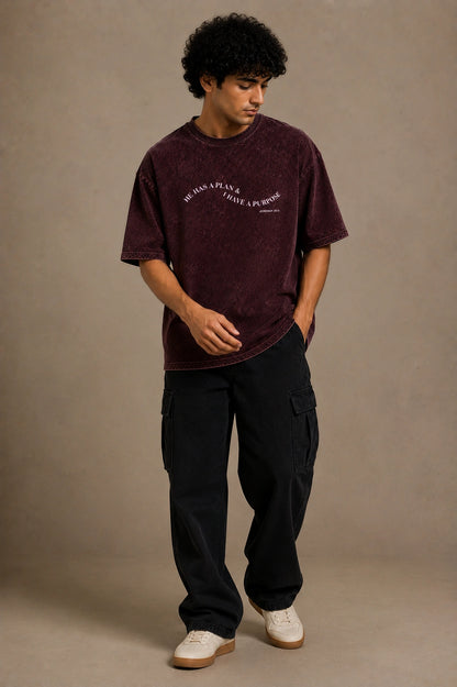 A man wearing a washed dark burgundy t-shirt with 'SANTO SEGUIN' printed on the chest, paired with black cargo pants and white sneakers, standing against a neutral beige studio backdrop.