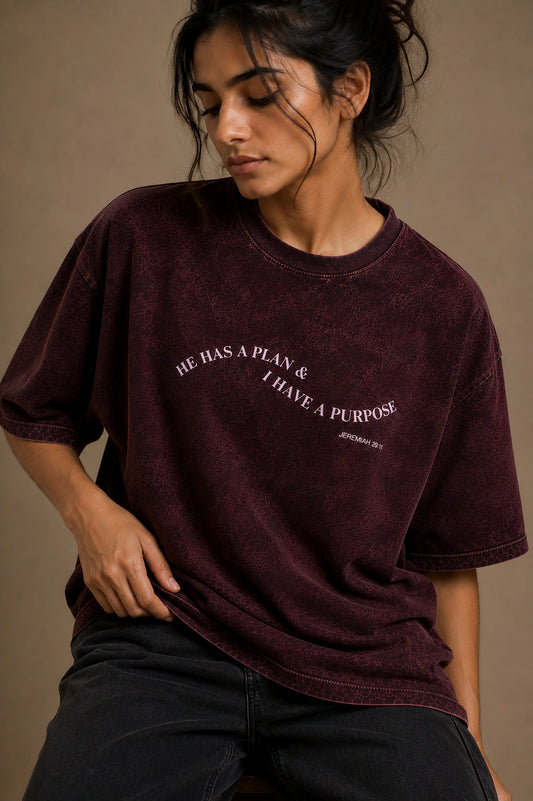 A woman wearing an oversized maroon mineral-wash t-shirt with the text 'HE HAS A PLAN.